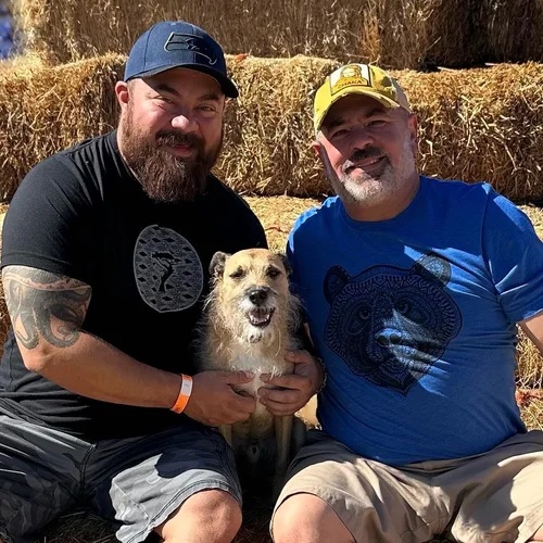 Chris and Adam with their dog Jasper, seated together in front of hay bales on a sunny day.